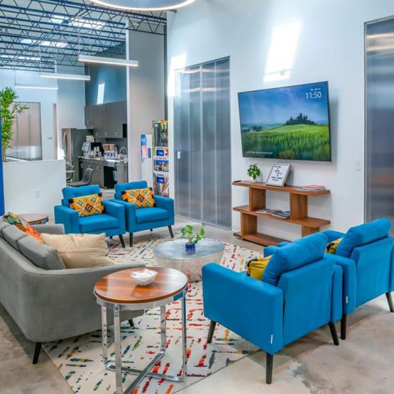 Modern lounge area with gray sofa, blue armchairs, geometric rug, metallic coffee table, and a wall-mounted TV displaying a scenic landscapeu00a0in a Central Texas coworking space
