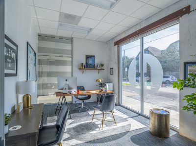 Bright modern office with a large window, wooden desk featuring a computer and lamp, stylish chairs, decorative art prints, and a view of a street with parked cars.