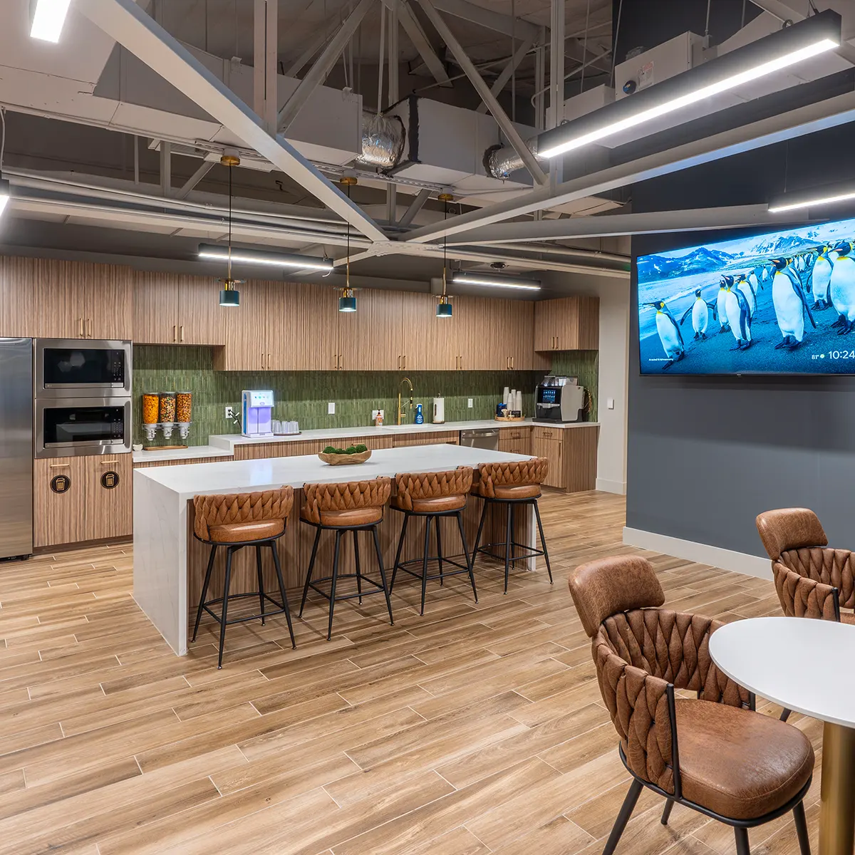 Modern office breakroom with a kitchen featuring wooden cabinets, green tile backsplash, a white island with leather bar stools, and a lounge area with tufted leather chairs and a TV displaying penguins in River Oaks, Houston, Texas